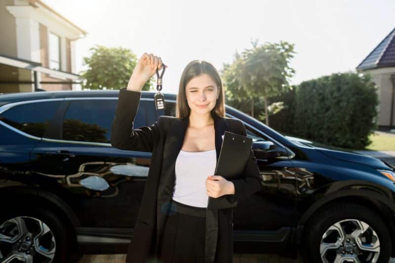 Buying or selling a car. Pretty young woman holding the keys standing in front of new black car