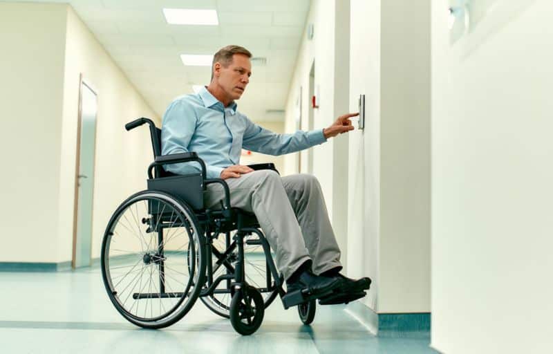 An elderly disabled man in a wheelchair presses the call button for the elevator in a modern clinic.