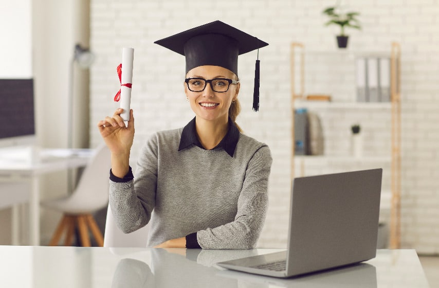Une femme dans une casquette de graduation tenant un diplôme devant un ordinateur portable.