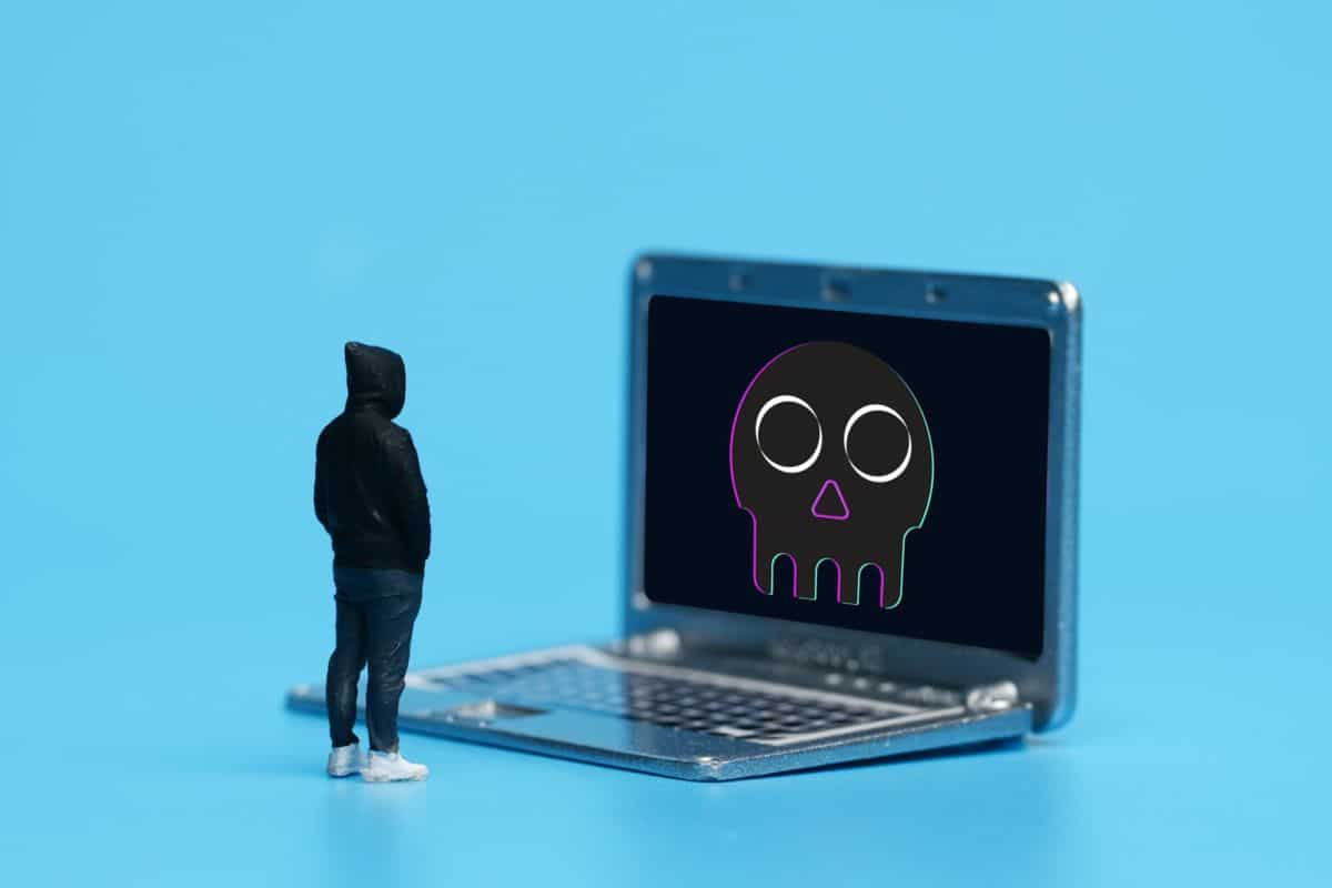 Une figurine miniature vêtue d'un sweat à capuche se tient face à un écran d'ordinateur portable affichant une icône de crâne de couleur néon, sur un fond bleu vif.