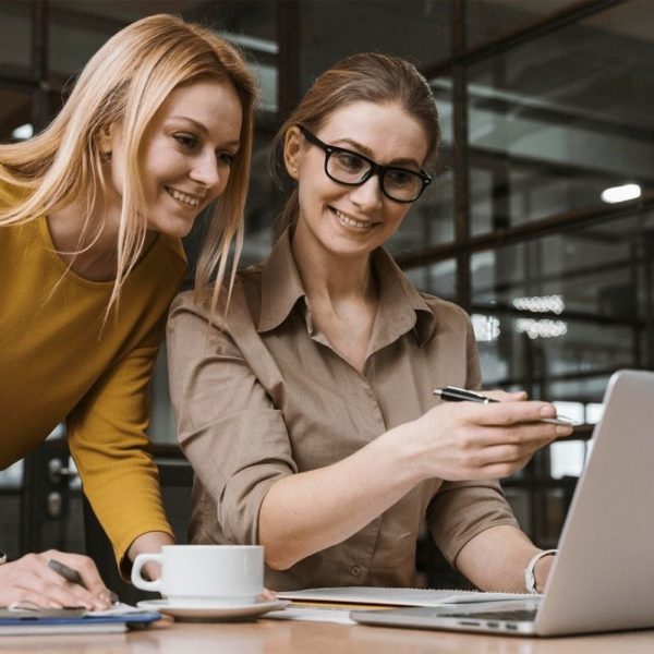 Deux Femmes En Tenue Professionnelle Décontractée Collaborent Autour D'un Ordinateur Portable Dans Un Bureau, L'une D'elles Pointant Vers L'écran Et Une Tasse De Café Visible.