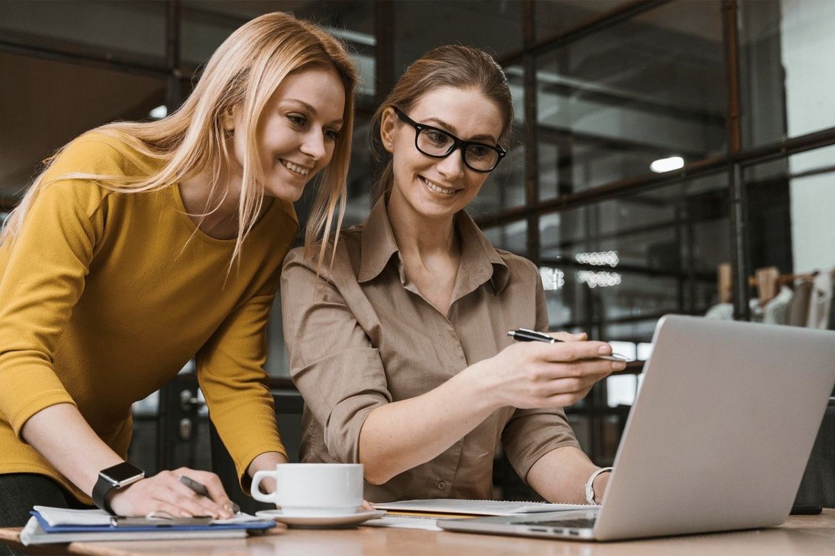 Deux Femmes En Tenue Professionnelle Décontractée Collaborent Autour D'un Ordinateur Portable Dans Un Bureau, L'une D'elles Pointant Vers L'écran Et Une Tasse De Café Visible.