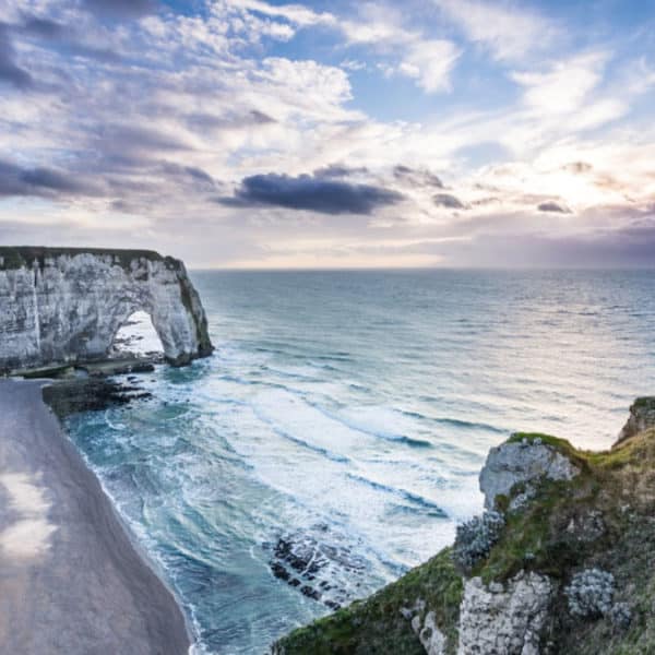 Une côte accidentée avec de hautes falaises blanches et une plage étroite qui s'étend le long du rivage, avec des vagues frappant les rochers et un ciel partiellement nuageux au-dessus.