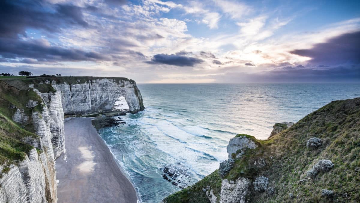 Une côte accidentée avec de hautes falaises blanches et une plage étroite qui s'étend le long du rivage, avec des vagues frappant les rochers et un ciel partiellement nuageux au-dessus.