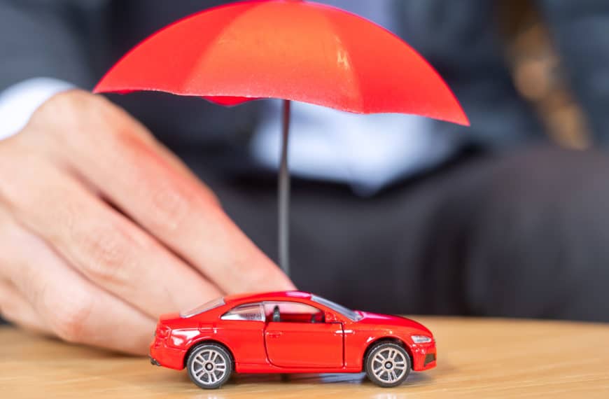 Une personne tient un petit parapluie rouge sur une voiture jouet rouge sur une surface en bois.