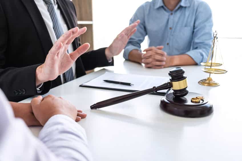 Une Personne En Costume Parle à Deux Autres Personnes Assises En Face D'un Bureau. Un Marteau, Une Balance Et Des Documents Sont Posés Sur Le Bureau.