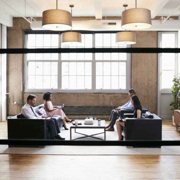 Quatre Personnes Sont Assises Sur Des Canapés Dans Un Bureau Moderne, Engagées Dans Une Discussion. La Pièce Est Dotée De Grandes Fenêtres, D'un Parquet En Bois Et De Plusieurs Luminaires. Une Imprimante Et Des Plantes Sont Visibles En Arrière Plan.