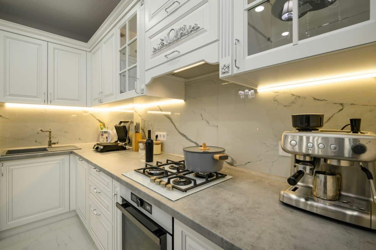 Cuisine Moderne Avec Armoires Blanches, Dosseret En Marbre Et Comptoirs Gris. Comprend Une Cuisinière Avec Une Casserole, Une Machine à Café Et Divers Appareils De Cuisine.