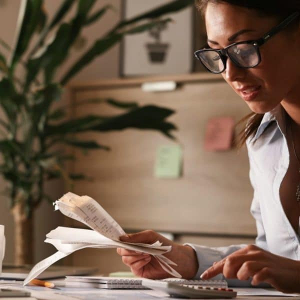 Une Femme Portant Des Lunettes Examine Des Reçus Et Utilise Une Calculatrice à Son Bureau. Un Ordinateur Portable, Un Cahier, Un Stylo Et Une Plante Se Trouvent également Sur Le Bureau.