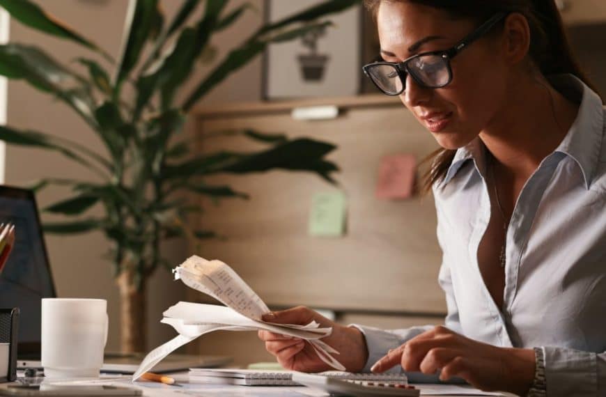 Une Femme Portant Des Lunettes Examine Des Reçus Et Utilise Une Calculatrice à Son Bureau. Un Ordinateur Portable, Un Cahier, Un Stylo Et Une Plante Se Trouvent également Sur Le Bureau.