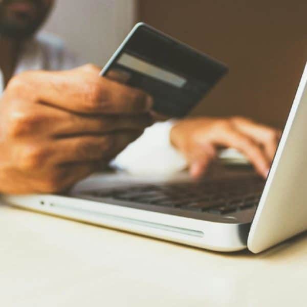Une Personne Tient Une Carte De Crédit Dans Sa Main Droite Tout En Utilisant Un Ordinateur Portable Placé Sur Un Bureau Avec Sa Main Gauche.