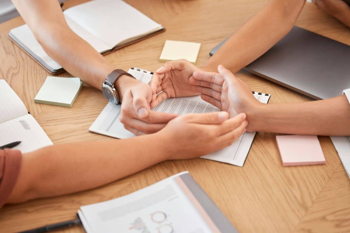 Quatre Personnes Sont Assises Autour D'une Table Avec Des Cahiers, Des Papiers Et Des Ordinateurs Portables, Tandis Que Deux D'entre Elles Se Tiennent La Main, Suggérant Un Travail D'équipe Ou Une Collaboration.
