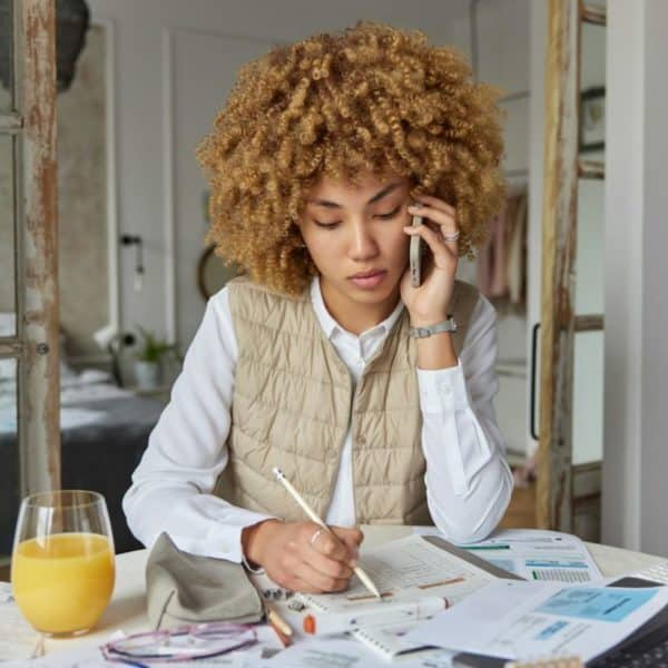 Une Personne Aux Cheveux Bouclés Parle Au Téléphone Tout En écrivant Dans Un Cahier. Un Verre De Jus D'orange, Des Verres Et Des Papiers Sont Posés Sur La Table.
