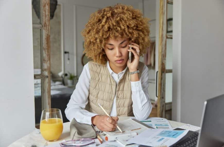 Une Personne Aux Cheveux Bouclés Parle Au Téléphone Tout En écrivant Dans Un Cahier. Un Verre De Jus D'orange, Des Verres Et Des Papiers Sont Posés Sur La Table.