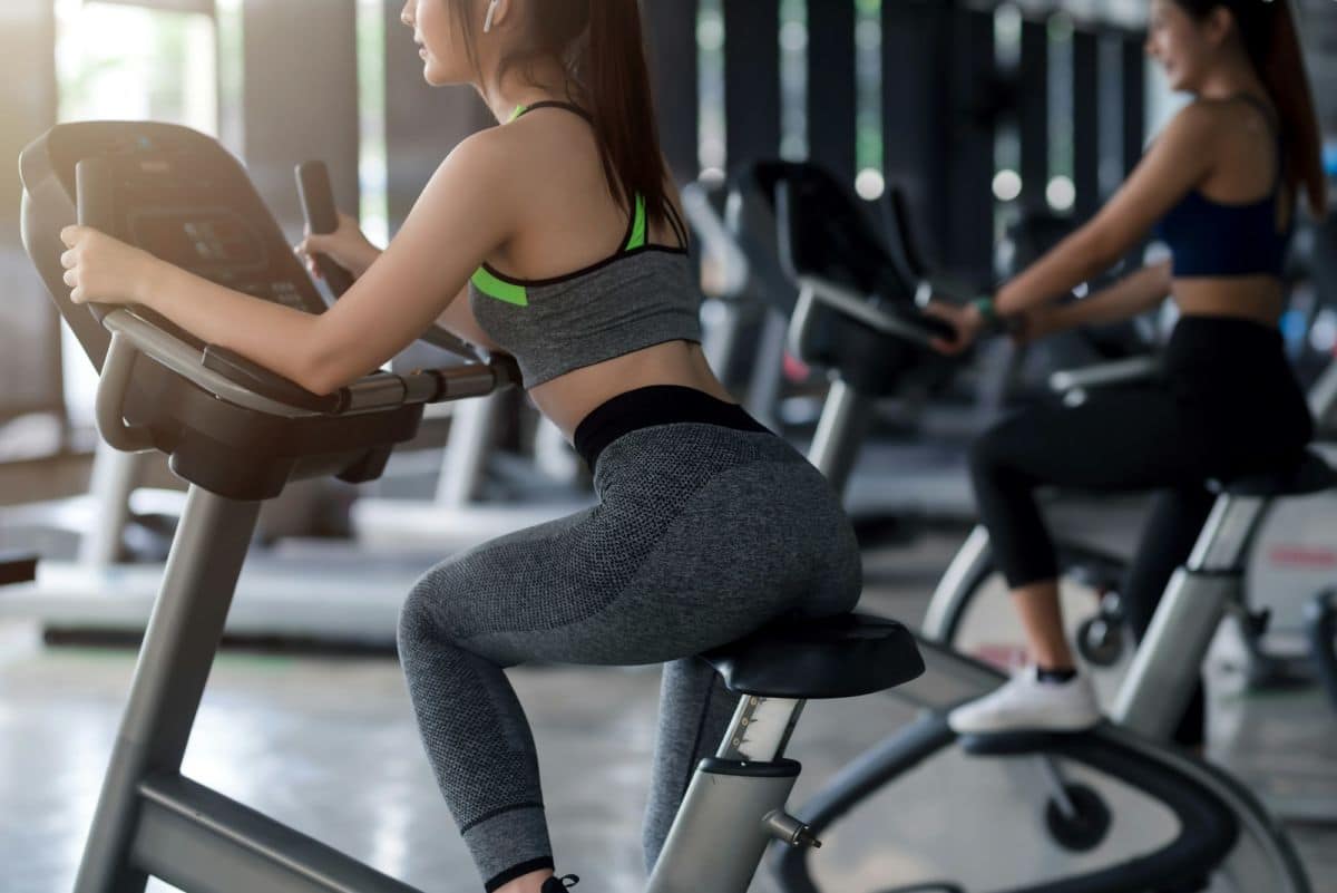 Deux personnes utilisent des vélos d'exercice stationnaires dans une salle de sport, l'accent étant mis sur la personne au premier plan portant une tenue d'entraînement grise.