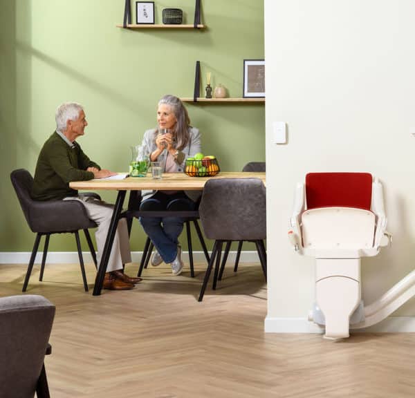 Deux Personnes âgées Sont Assises à Une Table Dans Un Salon Moderne Aux Murs Vert Clair. Un Monte Escalier Est Visible à Côté D'un Escalier Sur La Droite.
