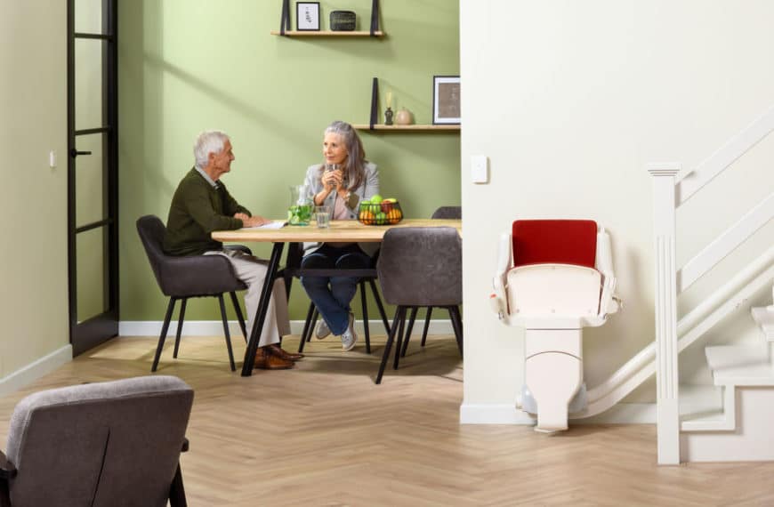 Deux Personnes âgées Sont Assises à Une Table Dans Un Salon Moderne Aux Murs Vert Clair. Un Monte Escalier Est Visible à Côté D'un Escalier Sur La Droite.