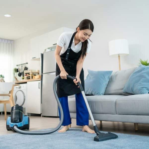 Une Personne Passe L'aspirateur Dans Un Salon Avec Un Aspirateur Bleu. La Pièce Dispose D'un Canapé Gris, D'une Lampe Et D'une Table à Manger.