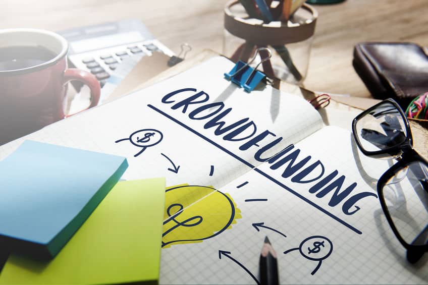 Un Carnet Avec L'inscription « Crowdfunding » écrite Dessus, Entouré De Notes Autocollantes Colorées, D'une Tasse à Café Et D'une Calculatrice Sur Une Table En Bois.