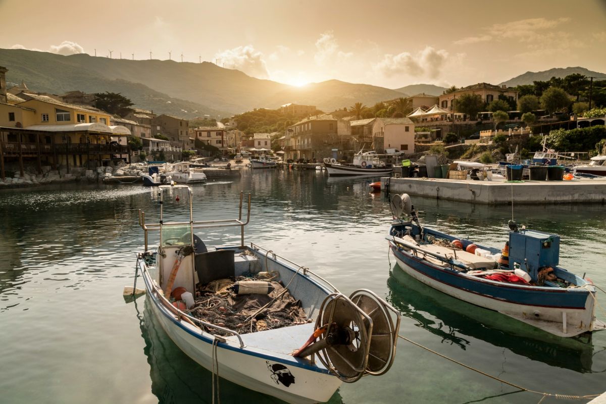 Deux petits bateaux de pêche amarrés dans un port calme au coucher du soleil en Corse, entourés de maisons et de montagnes en arrière-plan.
