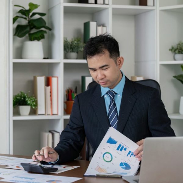 Un homme en costume examine des graphiques financiers à un bureau avec un ordinateur portable, une calculatrice et des documents. Des étagères avec des plantes sont visibles à l'arrière-plan.