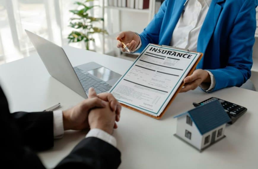 Deux Personnes Sont Assises à Un Bureau. L'une Tient Un Presse Papiers Avec Un Document D'assurance, Tandis Que L'autre écoute. Un Ordinateur Portable, Une Petite Maquette De Maison Et Une Calculatrice Sont Posés Sur La Table.
