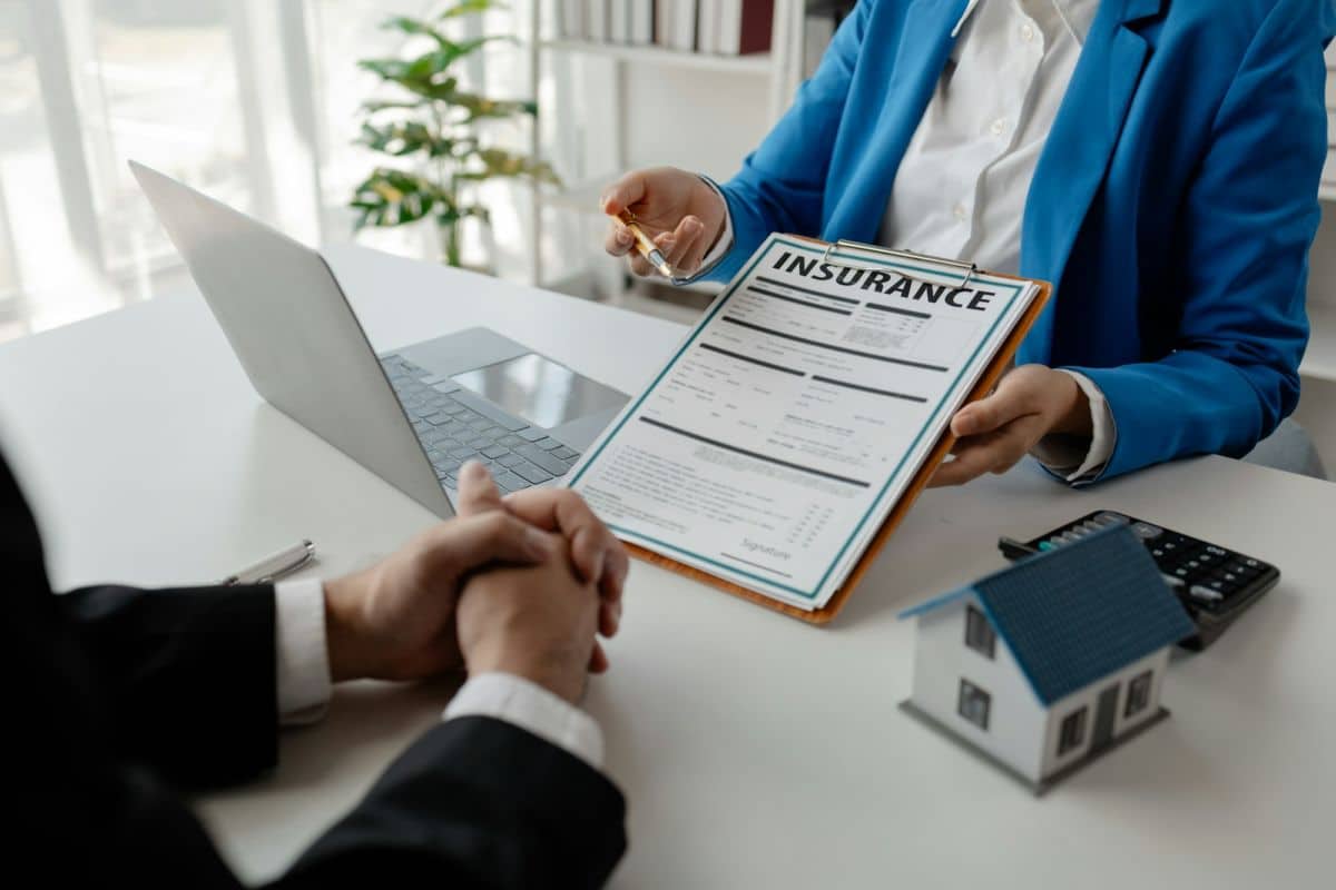 Deux Personnes Sont Assises à Un Bureau. L'une Tient Un Presse Papiers Avec Un Document D'assurance, Tandis Que L'autre écoute. Un Ordinateur Portable, Une Petite Maquette De Maison Et Une Calculatrice Sont Posés Sur La Table.