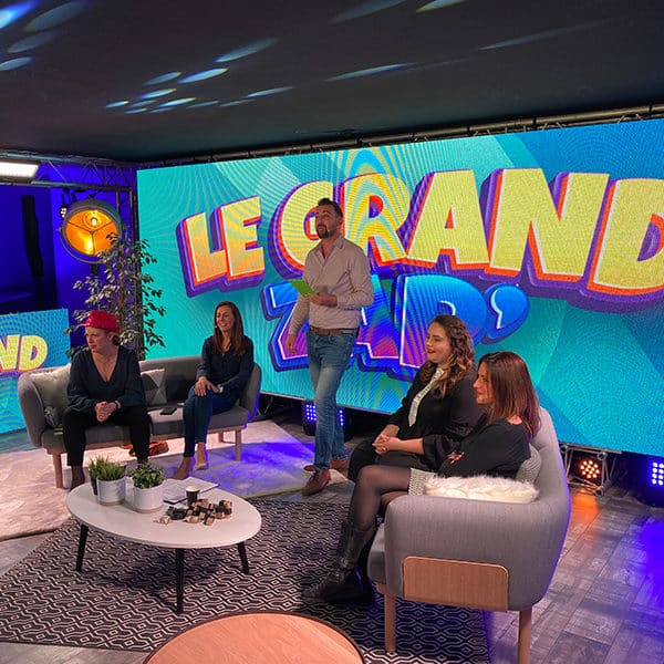 Cinq Personnes Sont Assises Et Debout Sur Un Plateau De Tournage Avec « Le Grand Zap » Projeté Sur Un Grand écran. L'installation Comprend Des Chaises, Une Table Et Des Plantes Décoratives.