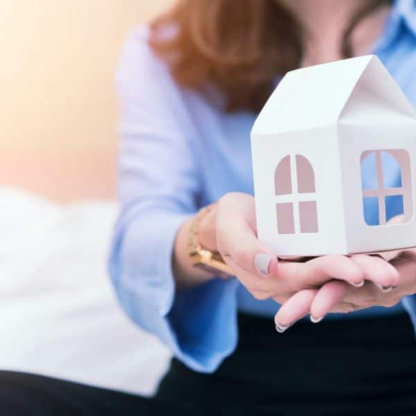 Une Femme En Chemise Bleue Tient Une Petite Maison Blanche Modèle Dans Ses Mains Ouvertes, Avec Un Arrière Plan Flou.
