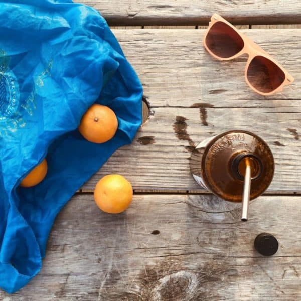 Un sac en tissu bleu avec des oranges, des lunettes de soleil roses et une bouteille en verre marron avec une paille sur une surface en bois.