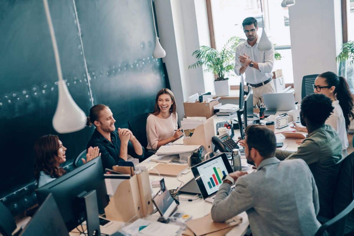 Un Groupe Diversifié De Collègues Est Assis Autour D'une Table Dans Un Bureau Et Participe à Une Discussion. Un Homme Se Tient Debout Et Parle Au Premier Plan, Tandis Que D'autres écoutent Et Applaudissent. Des Ordinateurs Et Des Graphiques Sont Visibles.