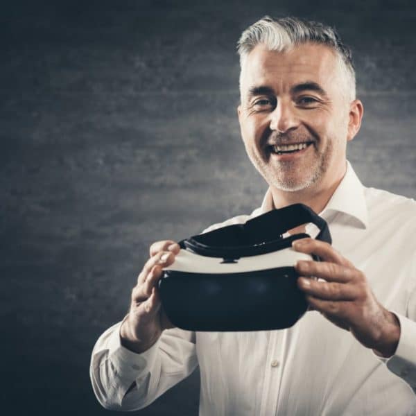 Un Homme En Chemise Blanche Tenant Un Casque Vr, Souriant Devant Un Fond Gris Foncé.