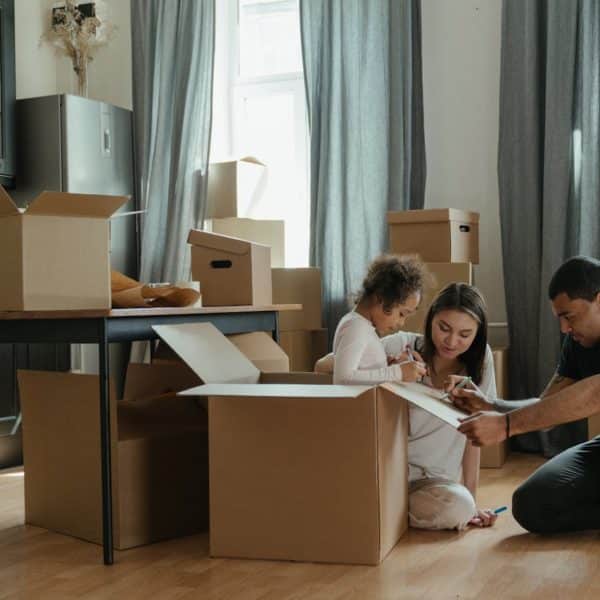 Une Famille Emballe Des Cartons Dans Un Salon, Avec Un Enfant Assis Dans Un Carton, Entouré De Cartons Ouverts Et Fermés.