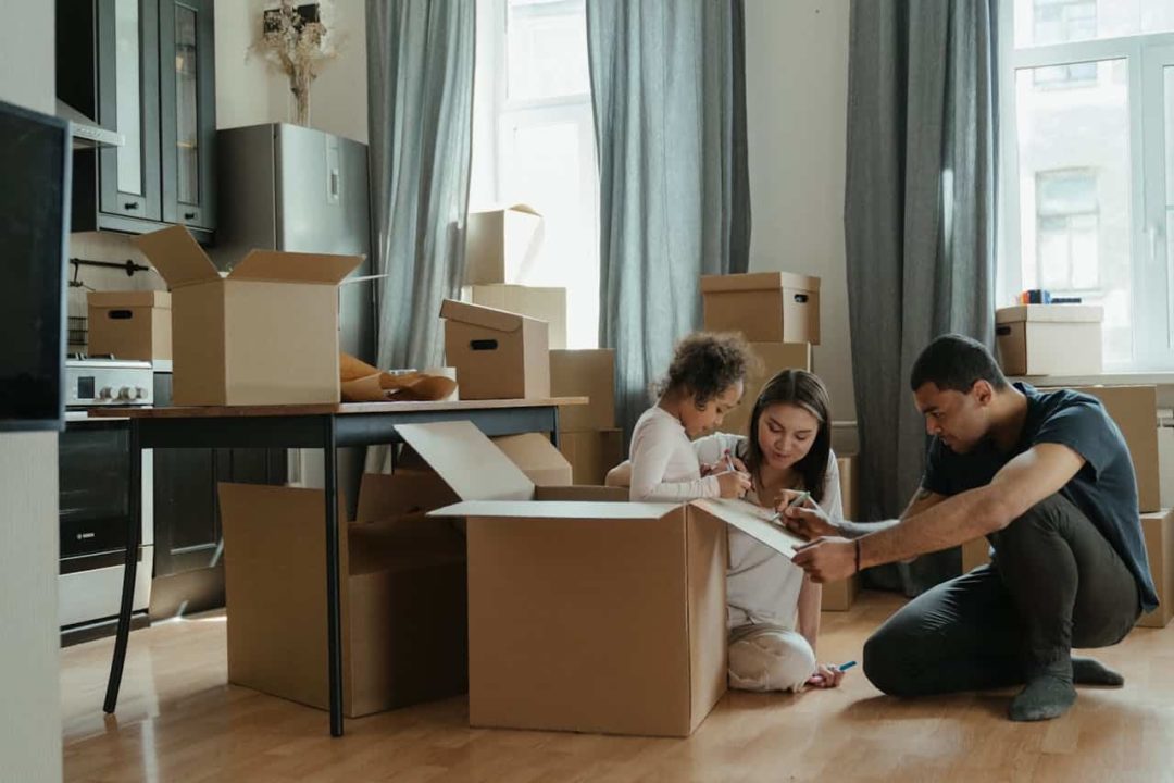 Une Famille Emballe Des Cartons Dans Un Salon, Avec Un Enfant Assis Dans Un Carton, Entouré De Cartons Ouverts Et Fermés.