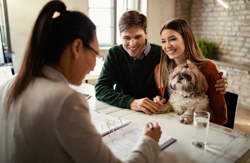 Un Couple Est Assis à Un Bureau Avec Un Chien, Interagissant Avec Une Femme Qui Tient Un Stylo Au Dessus D'un Dossier Ouvert.