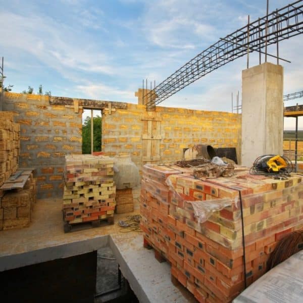 Des matériaux de construction empilés, notamment des briques et des tuiles, sont placés sur une structure partiellement construite. Des poutres en acier apparentes et une charpente ouverte sont visibles sous un ciel bleu.