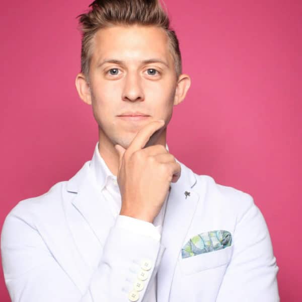 Un Homme En Costume Clair Avec Une Pochette Pose Sur Un Fond Rose Vif, Posant Pensivement Sa Main Sur Son Menton.