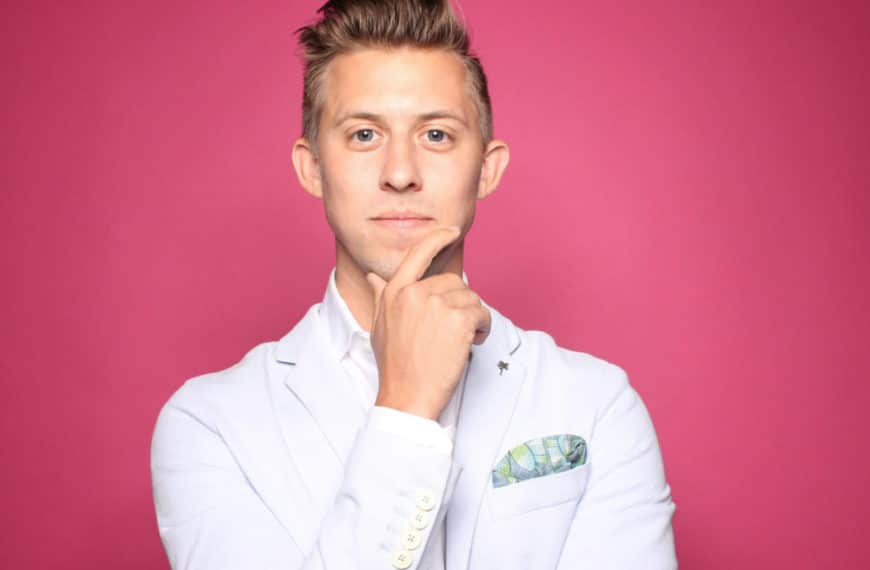 Un Homme En Costume Clair Avec Une Pochette Pose Sur Un Fond Rose Vif, Posant Pensivement Sa Main Sur Son Menton.