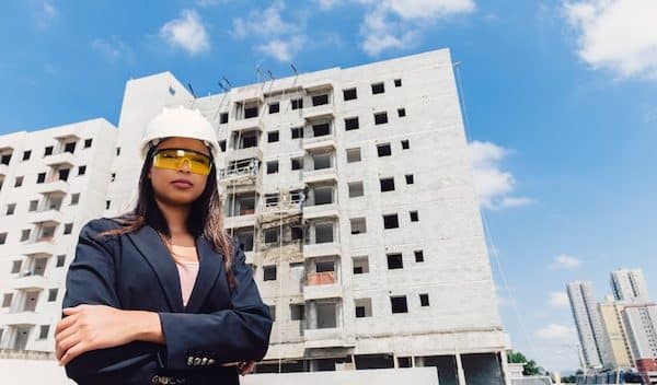 Une Femme Portant Un Casque Et Des Lunettes De Sécurité Se Tient Avec Confiance Devant Un Immeuble à Plusieurs étages En Construction.