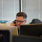 Un Homme Portant Des Lunettes Parle Au Téléphone Dans Un Bureau, Avec Des Ordinateurs Et Du Matériel De Bureau Autour De Lui.