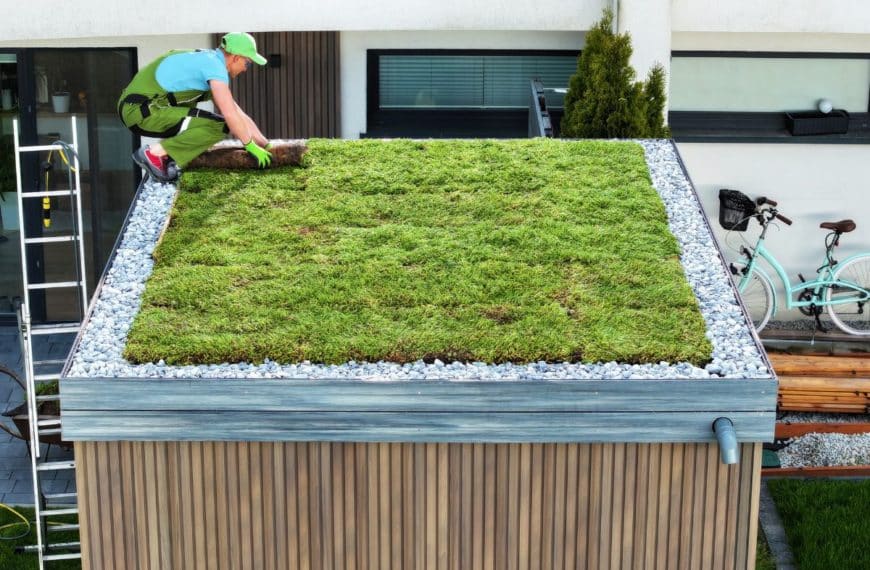 Une Personne En Tenue Verte Et Bleue Installe Du Gazon Sur Un Toit Végétalisé, Entouré De Bordures De Pierres. Un Vélo Est Visible à Proximité.