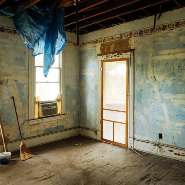 Ancienne Chambre En Cours De Rénovation, Poutres Apparentes Au Plafond, Murs Bleus écaillés, Deux Fenêtres, Un Climatiseur, Un Balai, Une Pelle à Poussière Et Une Bâche Bleue Suspendue Au Plafond.