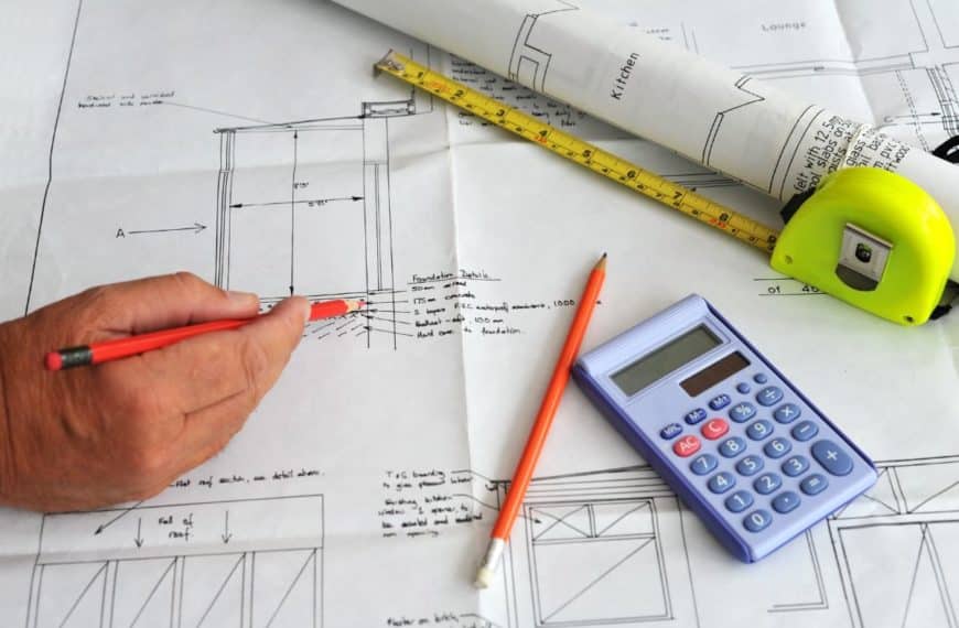 Dessin à La Main Sur Des Plans Architecturaux Avec Un Crayon Rouge, Planifiant Une Extension De Maison ; à Proximité Se Trouvent Une Calculatrice, Un Ruban à Mesurer Et Des Plans En Papier Roulé.