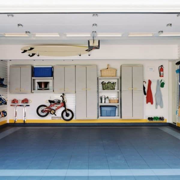Garage organisé avec placards de rangement, vélo, planche de surf et divers outils soigneusement rangés. Sol carrelé bleu et gris.