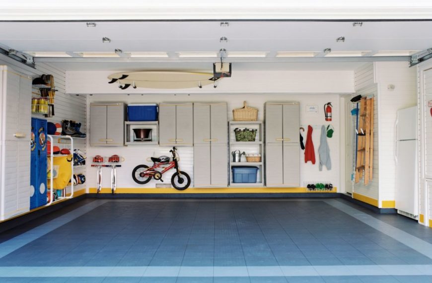 Garage organisé avec placards de rangement, vélo, planche de surf et divers outils soigneusement rangés. Sol carrelé bleu et gris.