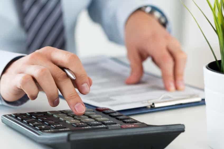 Une Personne Portant Un Costume Utilise Une Calculatrice Et Tient Un Stylo Au Dessus D'un Document Sur Un Presse Papiers ; Une Plante En Pot Est Visible à Proximité.