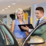 Un Homme En Costume Montre Une Tablette à Une Femme à Côté D'une Portière De Voiture Ouverte à L'intérieur D'un Concessionnaire Automobile.