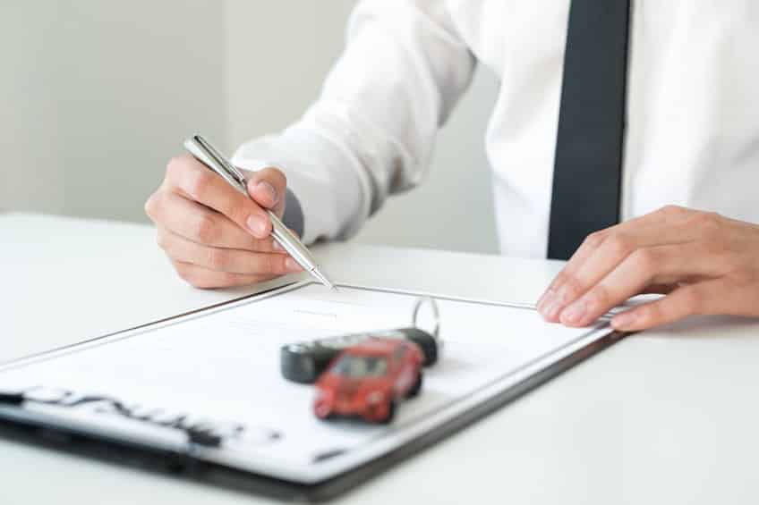 Une Personne En Tenue Professionnelle Signe Un Document Sur Un Presse Papiers, Avec Une Clé De Voiture Et Une Petite Voiture Placées Sur Le Dessus Du Papier.