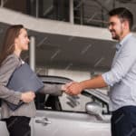Une Femme Tenant Un Dossier Et Un Homme Se Serrent La Main Devant Une Voiture Dans La Salle D'exposition D'un Concessionnaire.