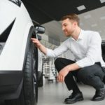 Un homme vêtu d'une chemise blanche et d'un pantalon foncé s'accroupit pour inspecter le pneu avant d'une voiture blanche à l'intérieur d'une salle d'exposition.